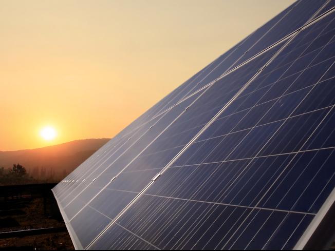 A closeup of a solar panel at an angle. In the distance, the sun sets over the hills